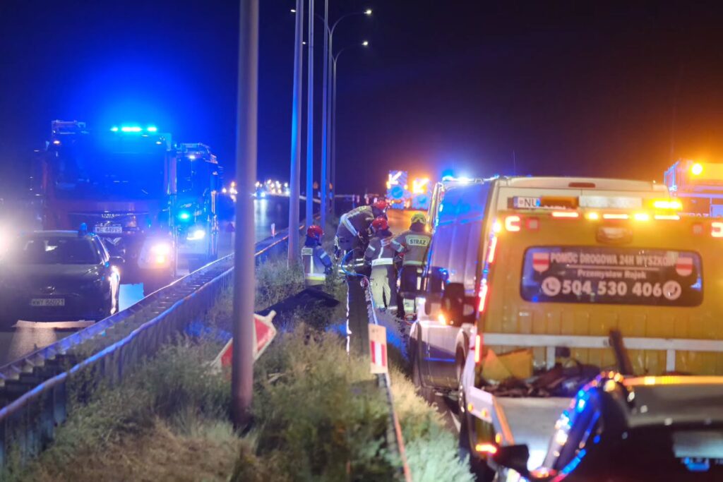 Grupa strażaków udziela pomocy poszkodowanemu na trasie S8, w tle widać pojazdy ratunkowe z włączonymi sygnałami świetlnymi.
