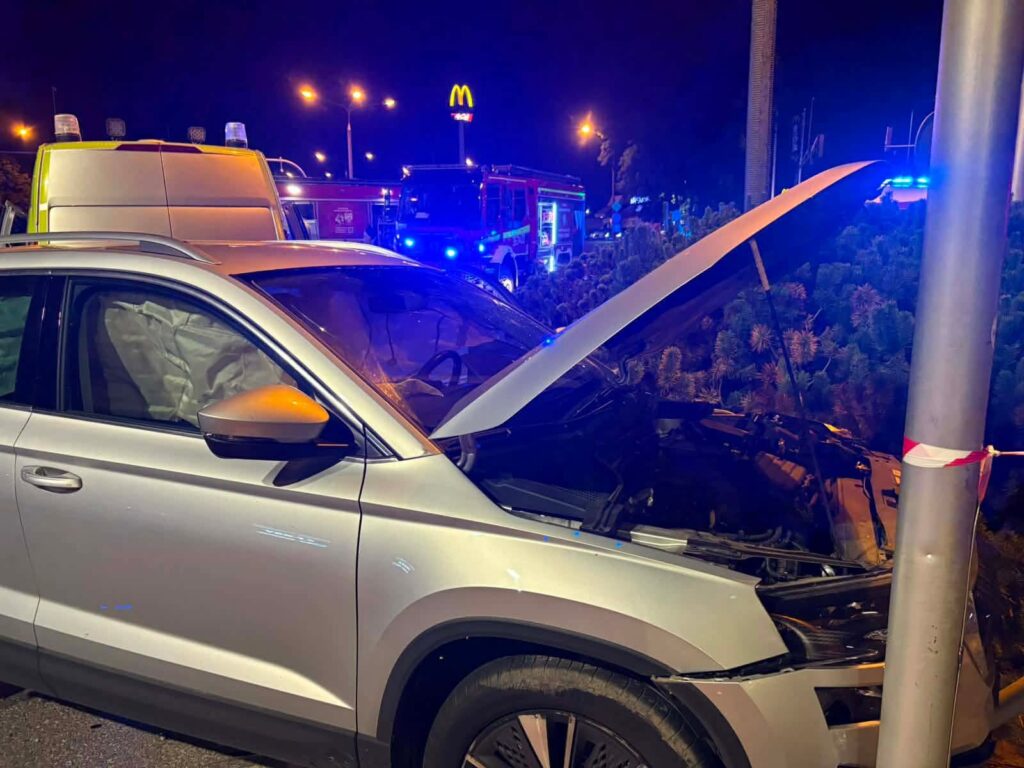 Srebrny SUV z otwartą maską uderzył w latarnię, w tle widać pojazdy straży pożarnej i migające światła.