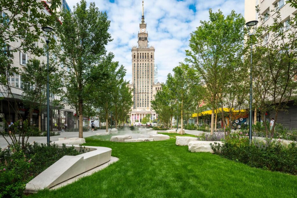 Zielona przestrzeń z trawnikiem i drzewami, w tle wieżowiec z zegarem, na ulicy z nowoczesnymi elementami architektonicznymi.