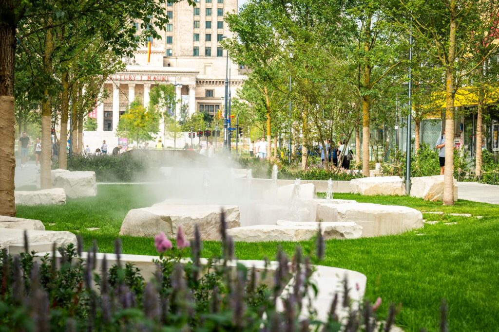 Nowoczesny park z fontannami i zielonym trawnikiem, otoczony drzewami, w tle widoczny budynek z kolumnami.