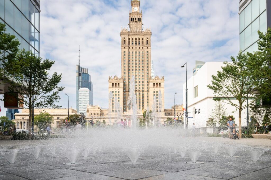 Fontanna tryska wodą na nowo wyremontowanej ulicy, w tle widoczny Pałac Kultury i Nauki oraz nowoczesne budynki.