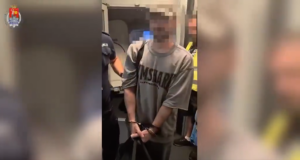 Mężczyzna w szarej koszulce, skuty w kajdankach, stoi w towarzystwie policjantów na pokładzie pojazdu transportowego.