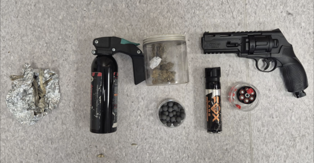 Na szarej powierzchni leżą: czarny pistolet, pojemnik z kulkami, gaśnica, słoik z substancją roślinną oraz ampułka.