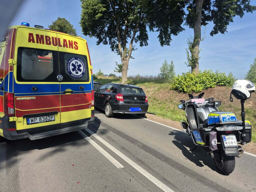 Karetka pogotowia oraz policyjny motocykl stoją na drodze obok czarnego samochodu osobowego.