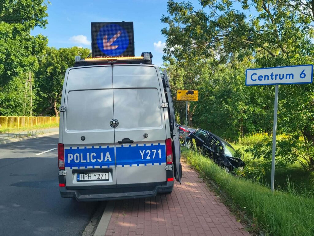 Policyjny samochód zaparkowany na poboczu drogi, obok znaku kierunkowym z napisem "Centrum 6", w tle uszkodzony pojazd.