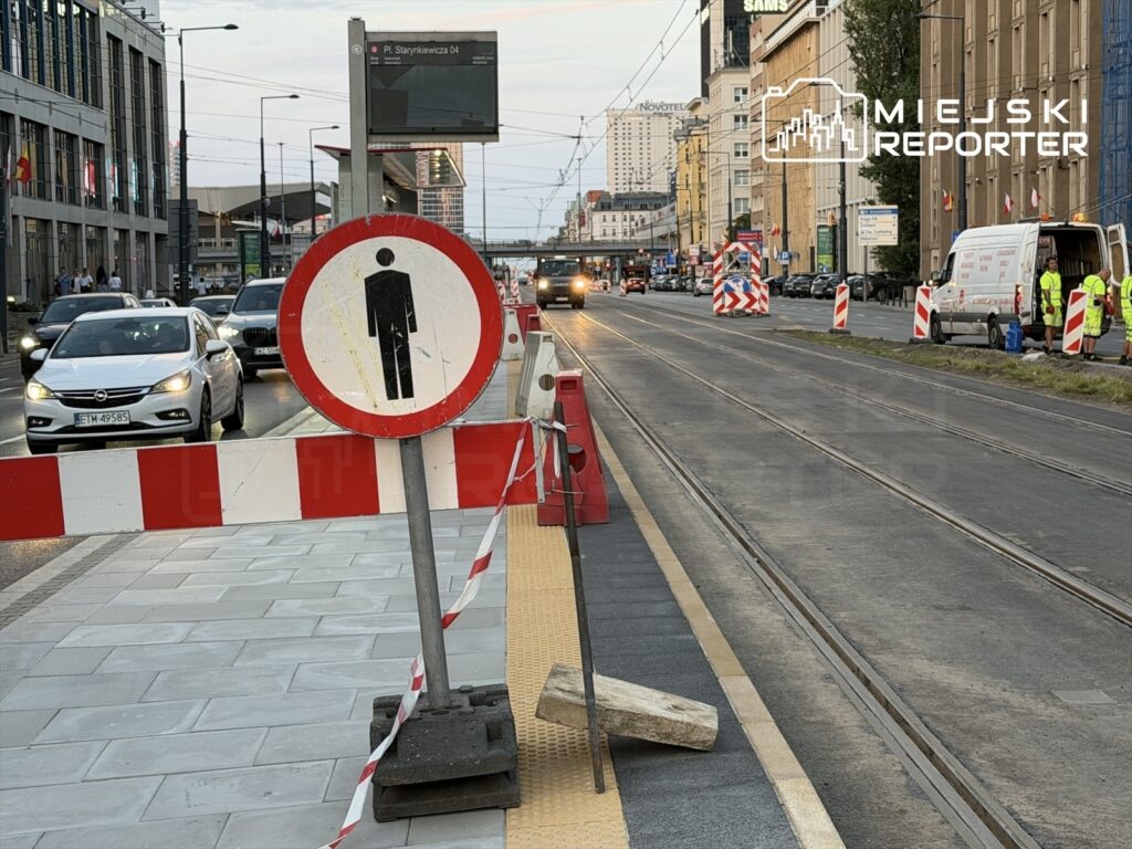 W ruchliwej ulicy znajdują się znaki drogowe oraz barierki budowlane, a w tle widać samochody i przechodniów.