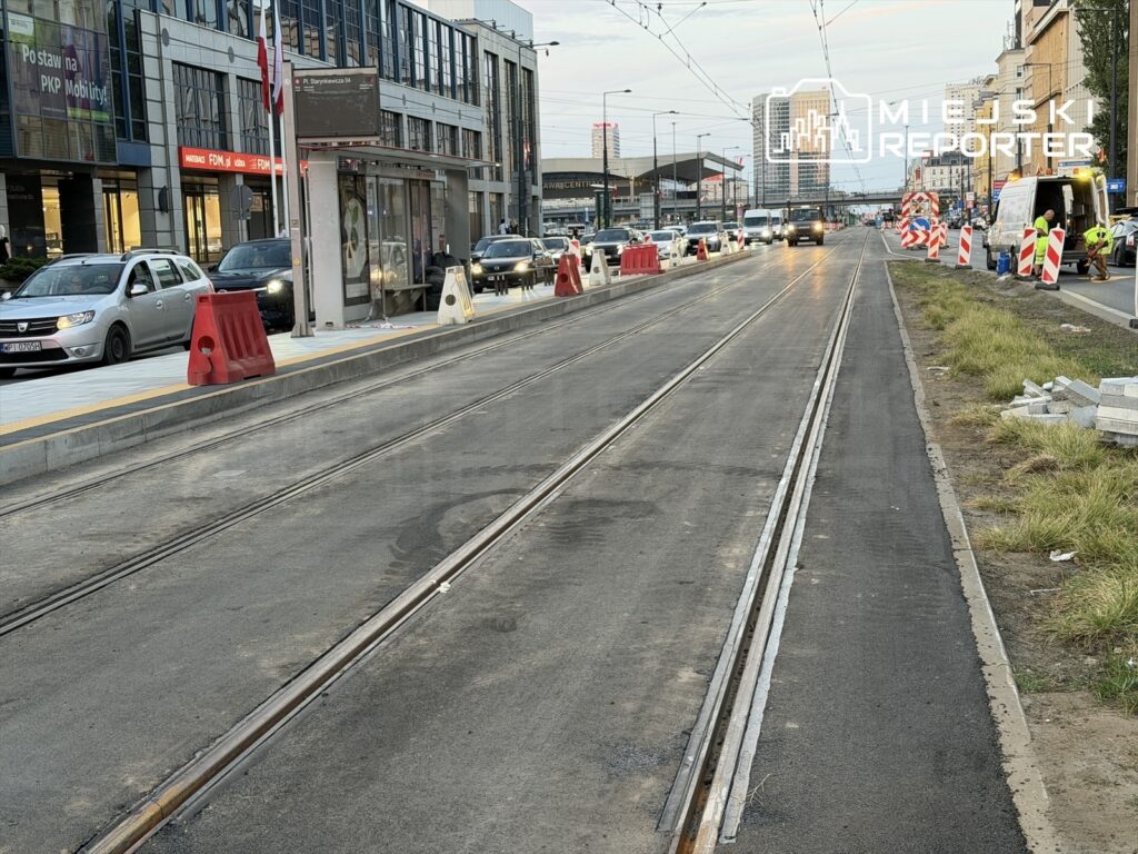 Ruch uliczny w pobliżu budowy torowiska, z samochodami stojącymi w korku i pracownikami w kamizelkach odblaskowych.