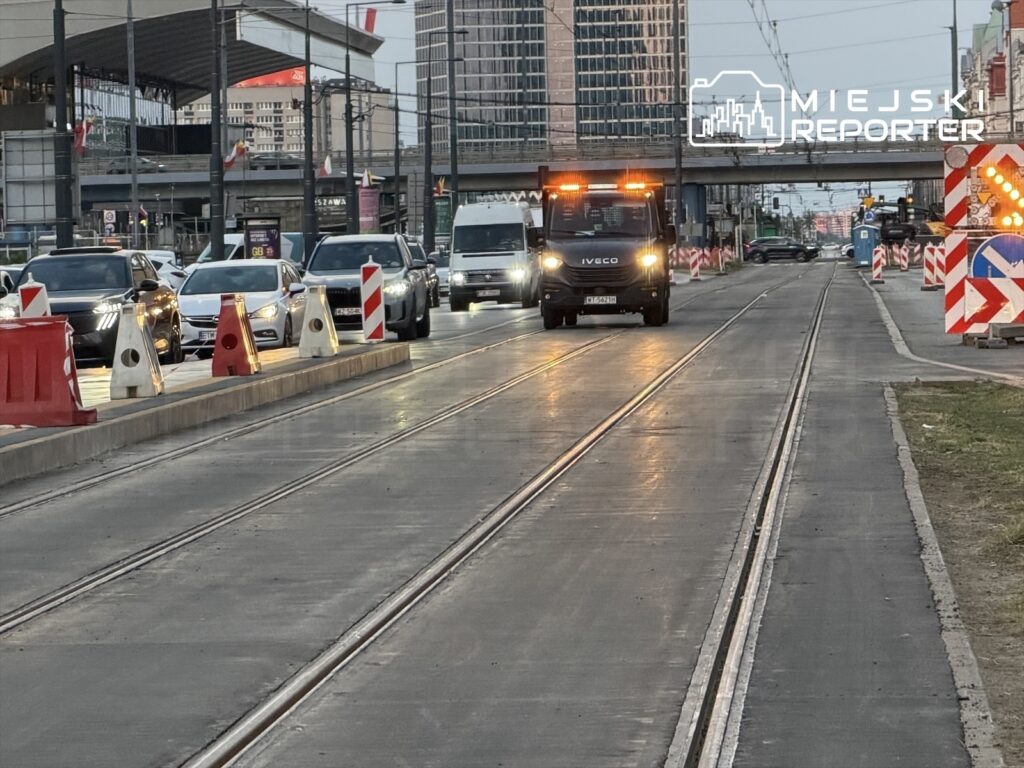 Pojazd służbowy z pomarańczowymi światłami przejeżdża obok samochodów w ruchu, na drodze z torami tramwajowymi.