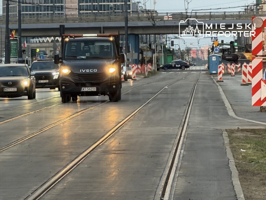Ciężarówka Iveco porusza się po ulicy, obok innych samochodów, w otoczeniu robót drogowych.