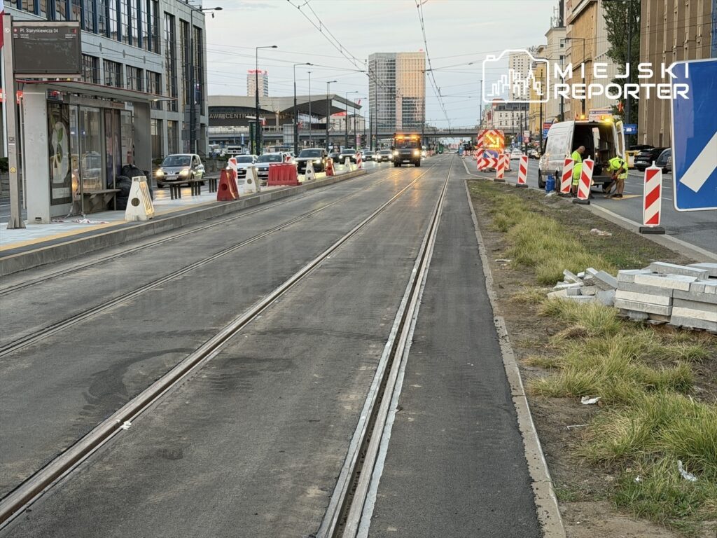 Robotnicy pracujący przy budowie torowiska tramwajowego na ruchliwej ulicy, otoczeni barierkami drogowymi.