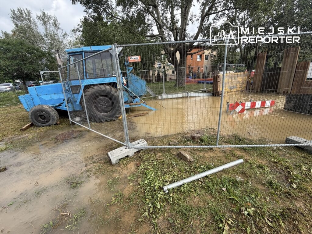 Niebieski traktorek stojący w zalanym terenie, otoczony ogrodzeniem budowlanym, obok widoczne znaki ostrzegawcze.