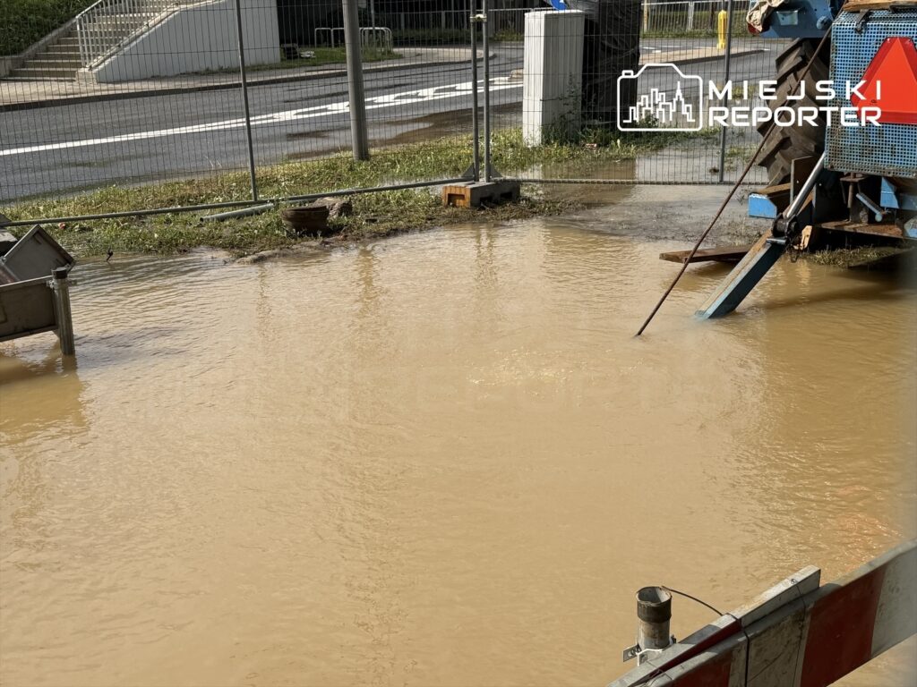 Zalany teren budowy z brązową wodą, obok widoczny sprzęt budowlany i ogrodzenie.