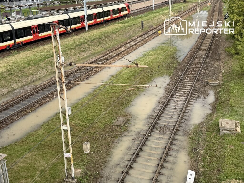Tor kolejowy z widocznymi kałużami wody, obok pociąg stojący na stacji. Zielona trawa w tle.