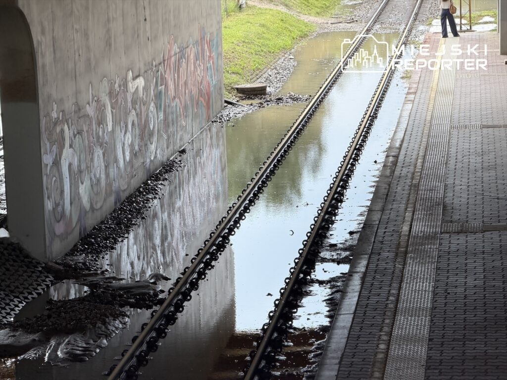 Woda stojąca na torach kolejowych obok graffiti na betonowej ścianie, w tle widać peron.