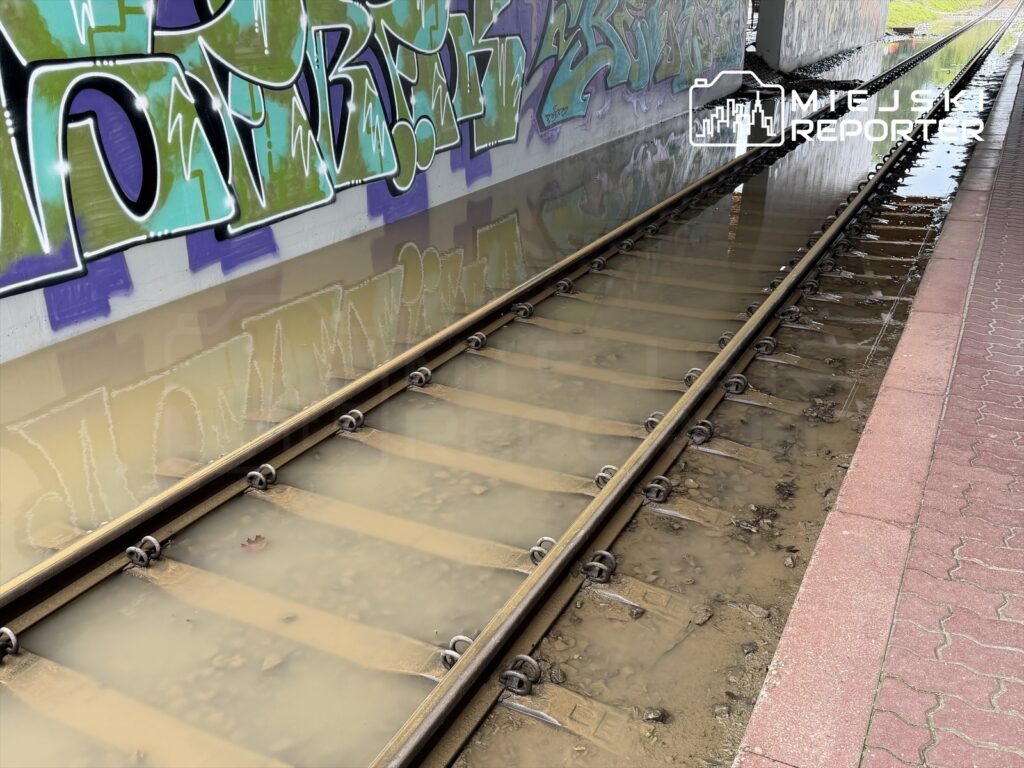 Zanurzone w wodzie tory kolejowe pod graffiti, z widoczną wodą i zanieczyszczeniami na torach.