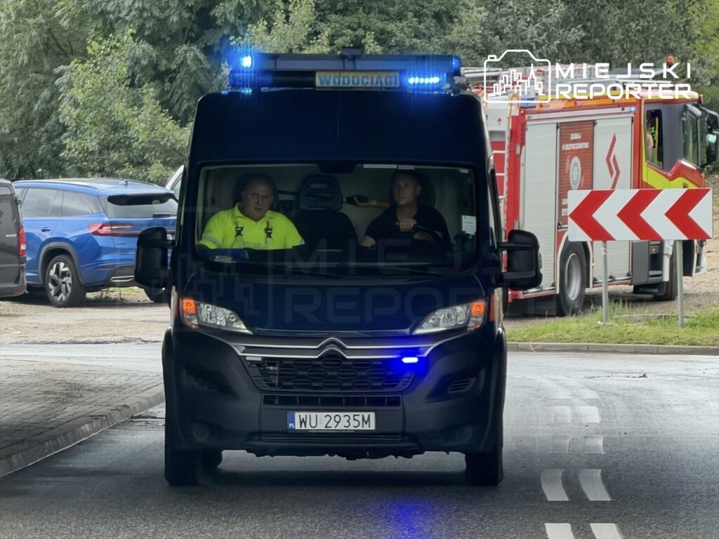 Wóz strażacki z włączonymi sygnalizatorami świetlnymi przejeżdża przez skrzyżowanie, obok samochodów osobowych.