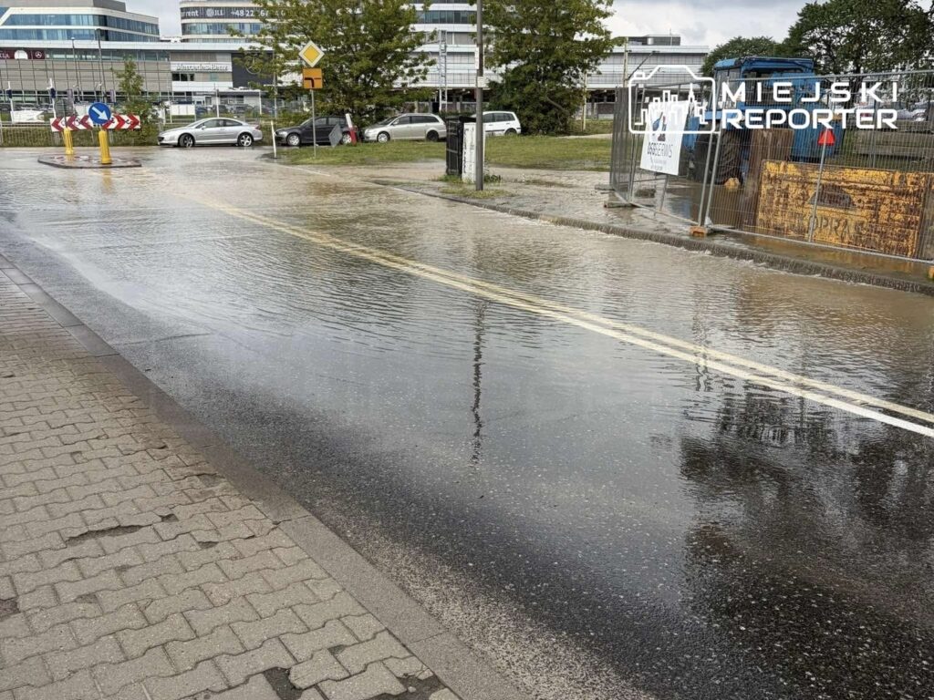 Zalana ulica z widocznymi kałużami wody, obok ronda i ogrodzenia budowy, w tle nowoczesne biurowce.