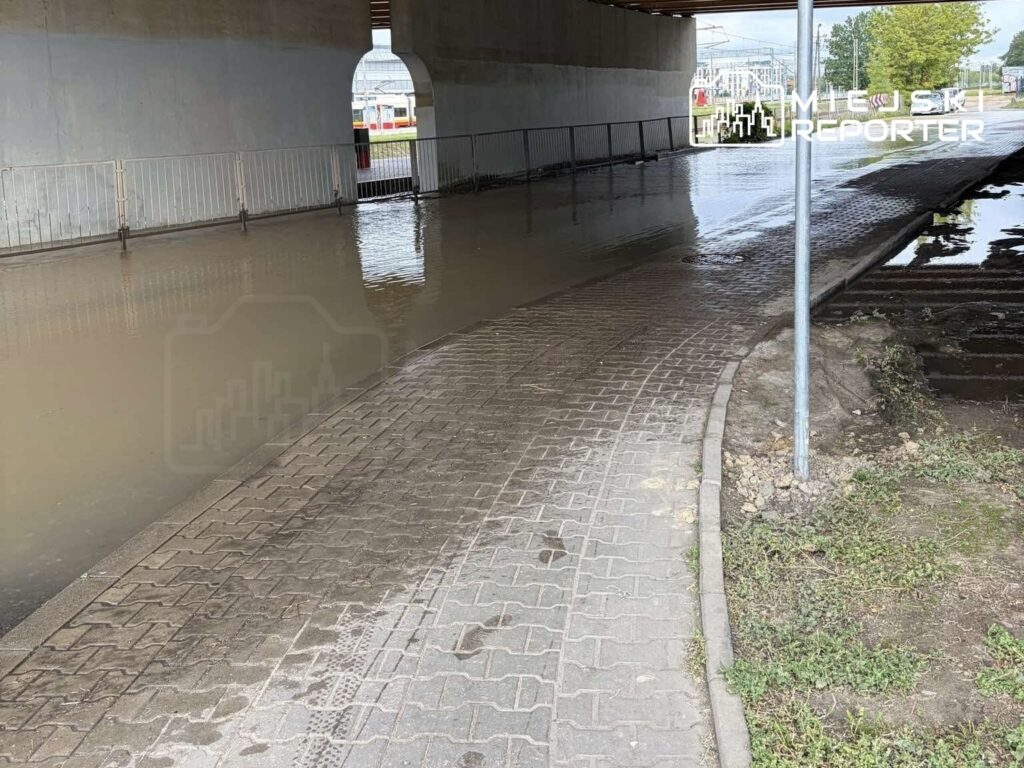 Zalana droga pod wiaduktem, z widoczną wodą i przeszklonymi budynkami w tle. Pojazdy nie przejeżdżają.
