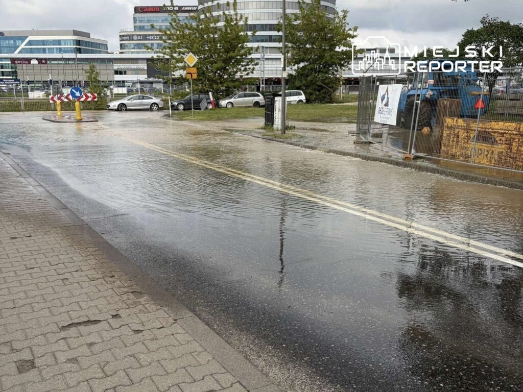 Zalany odcinek drogi z widocznymi kałużami wody, obok modernizowanej przestrzeni z ogrodzeniem budowlanym.