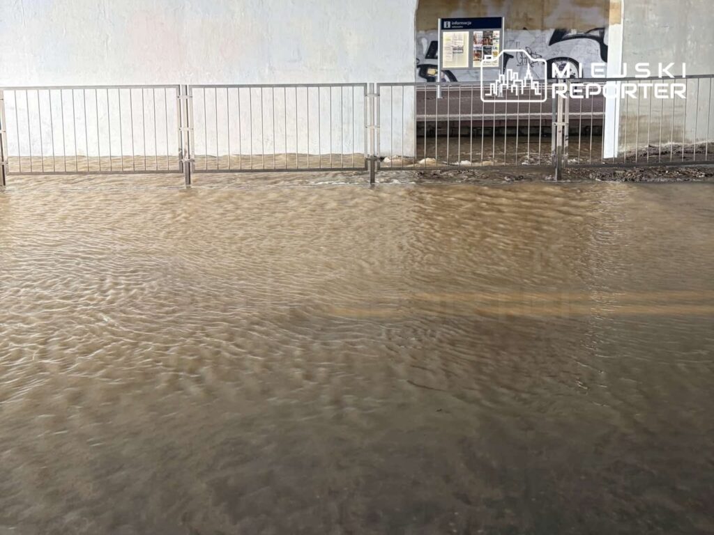 Zalany teren pod wiaduktem, z widoczną barierką, woda tworzy brązową taflę, zatapiając okolice przystanku.
