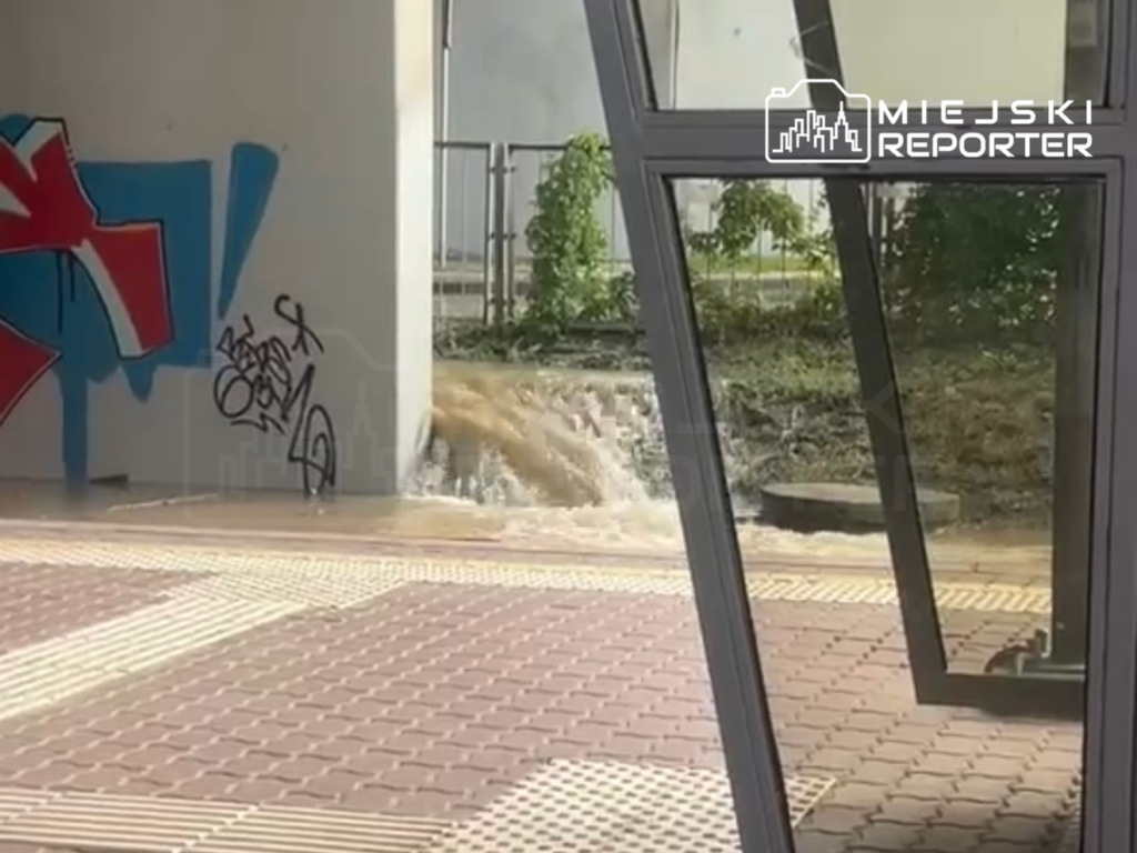 Woda wypływa z uszkodzonej rury w pobliżu graffiti na ścianie, tworząc kałużę na chodniku.