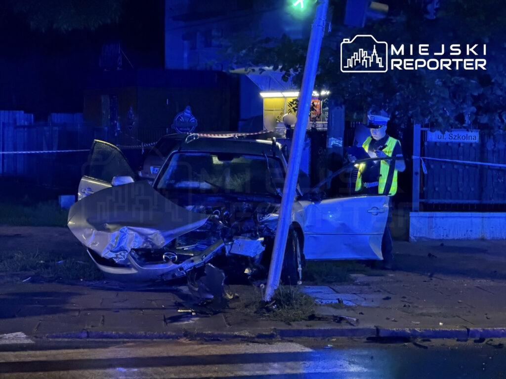 Uszkodzony samochód osobowy z wgniecionym przodem stoi obok słupa energetycznego, przy którym pracuje policjant.