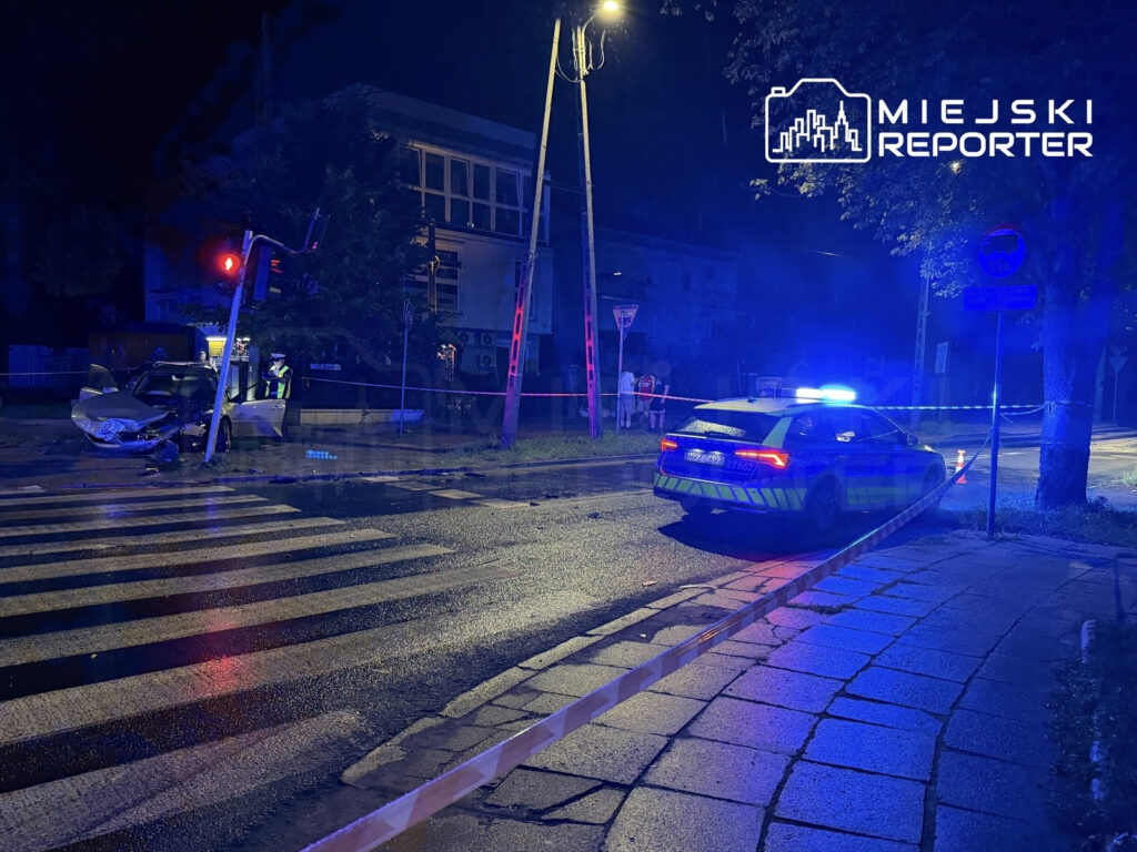 W nocy, w deszczowej scenerii, policjant bada uszkodzony samochód przy czerwonym sygnalizatorze na ulicy.