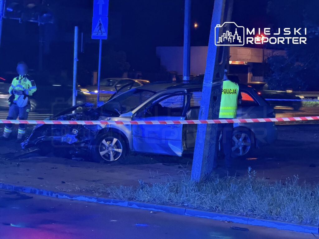 Pojazd osobowy z uszkodzoną przednią częścią stoi przy drodze, obok policjant w kamizelce odblaskowej prowadzi czynności na miejscu zdarzenia.