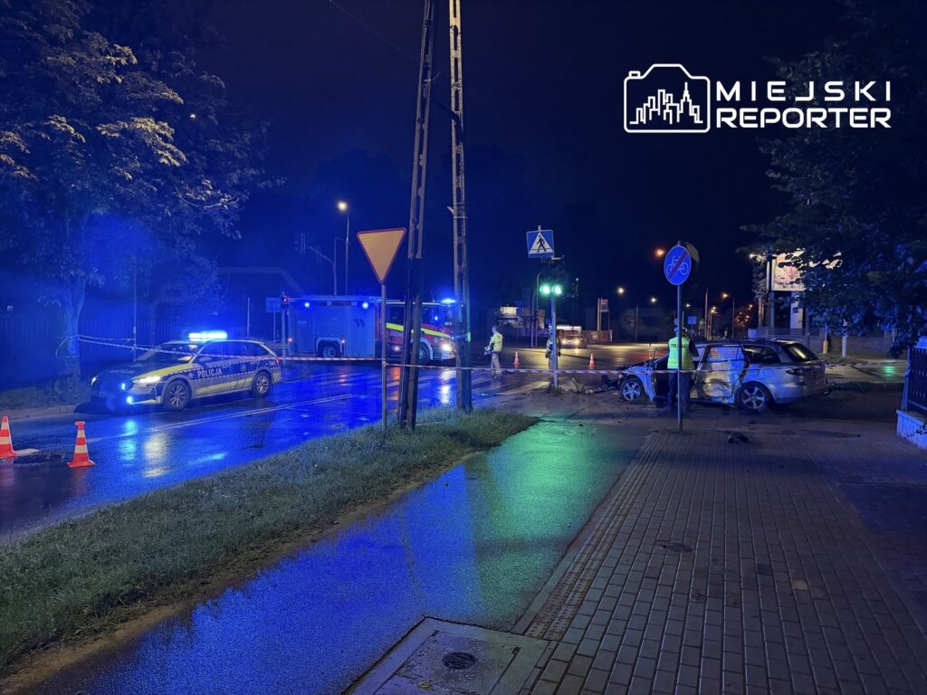 Policyjny radiowóz z włączonymi sygnałami świetlnymi na oświetlonej ulicy po nocnym wypadku, obok uszkodzonego samochodu.