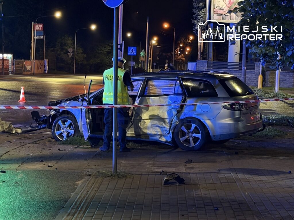 Policjant w kamizelce odblaskowej bada uszkodzony samochód osobowy przy drodze, otoczony taśmą zabezpieczającą.