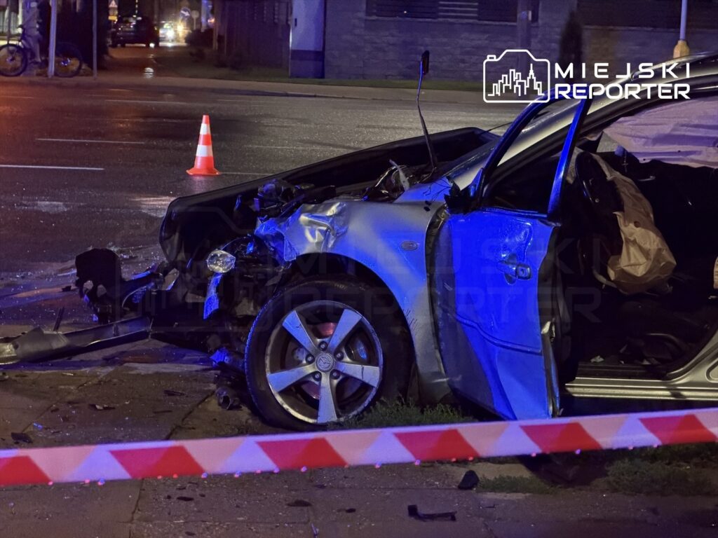 Zniszczony samochód osobowy z otwartymi drzwiami na poboczu drogi, otoczony taśmą zabezpieczającą.