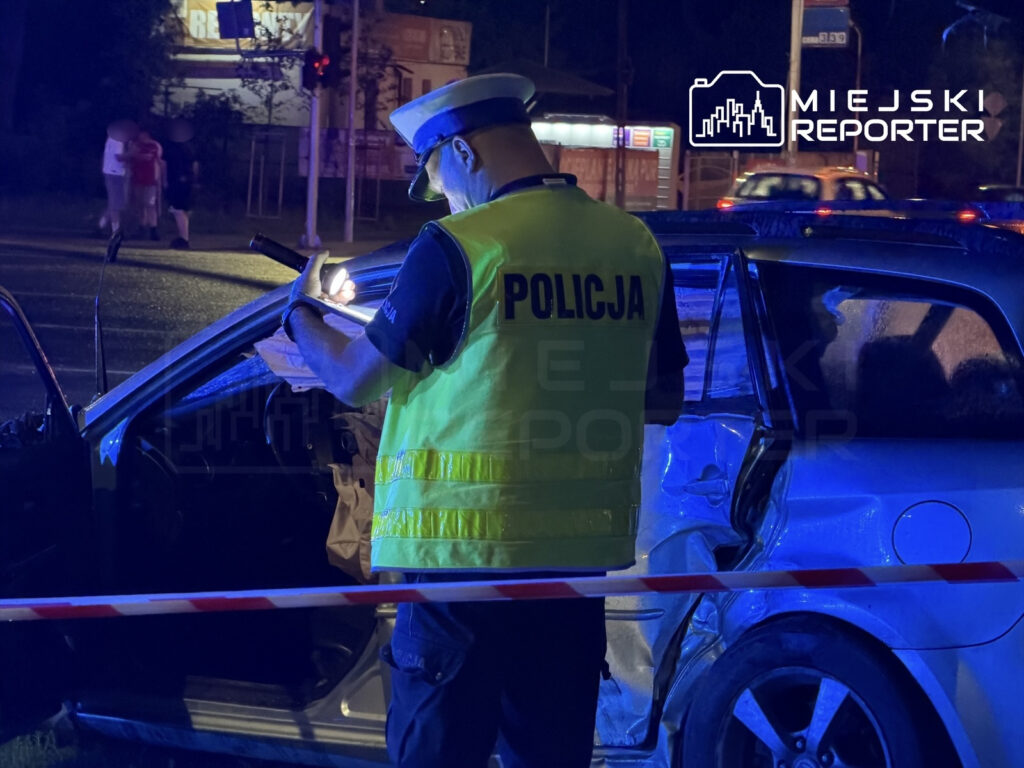 Policjant w kamizelce odblaskowej prowadzi oględziny uszkodzonego samochodu w nocy, przy oświetleniu policyjnym.