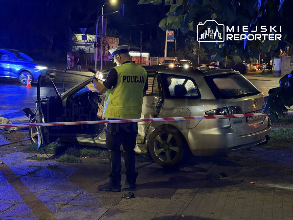 Policjant w kamizelce odblaskowej bada uszkodzone auto zaparkowane przy ulicy, otoczone taśmą ostrzegawczą.