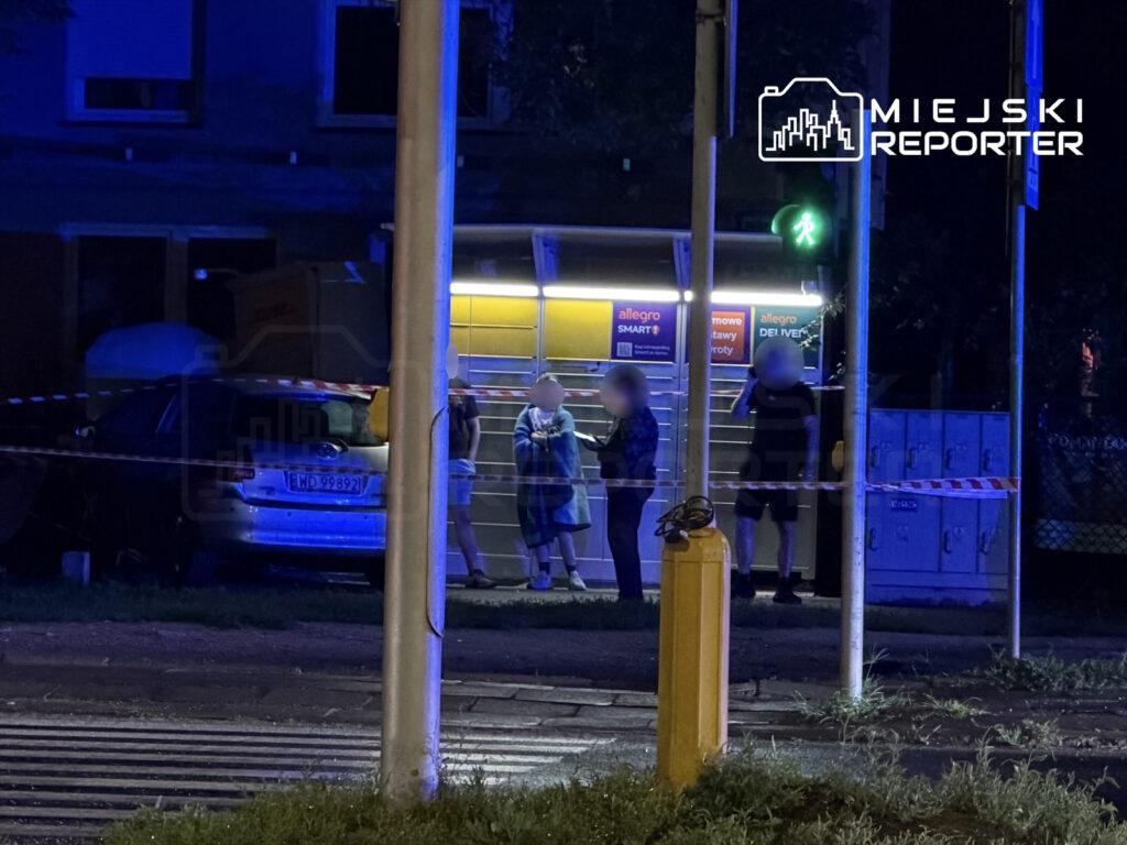 Grupa osób rozmawia przy oświetlonym kiosku, obok zaparkowanego samochodu, w nocy.