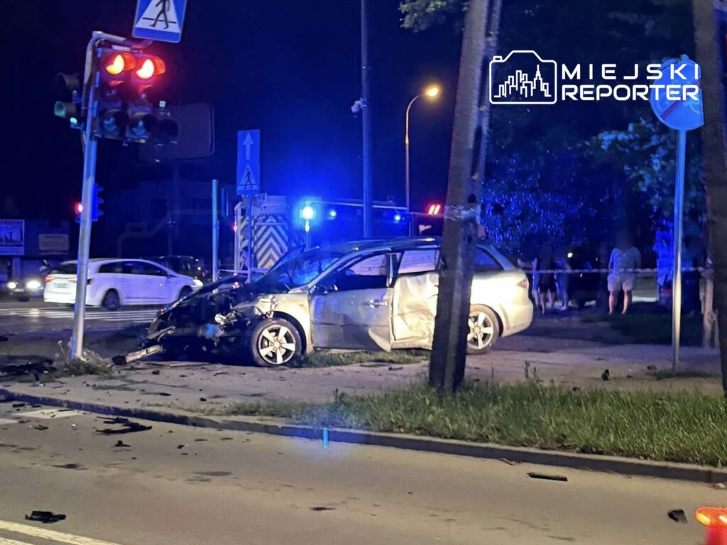 Samochód osobowy zniszczony w wyniku wypadku, stoi na chodniku obok sygnalizacji świetlnej w nocy. W tle widać radiowóz i grupę osób.
