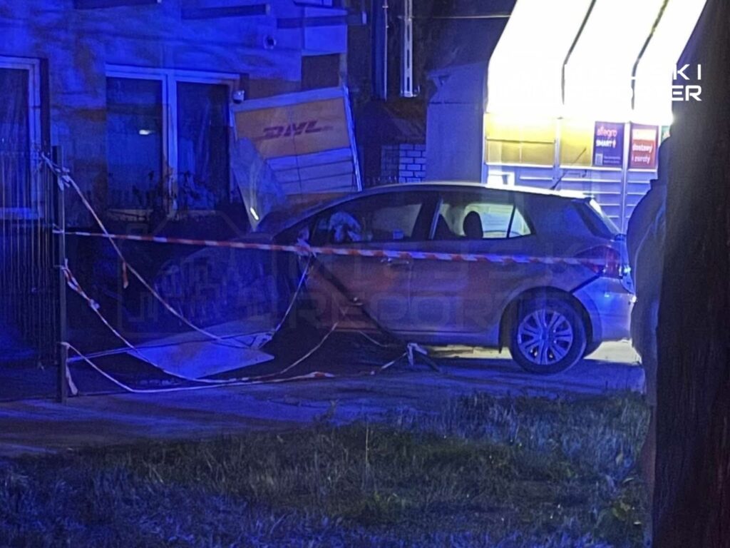 Samochód osobowy stoi przy ogrodzeniu, częściowo zasłonięty przez niebieskie światła, obok widoczna uszkodzona furgonetka.