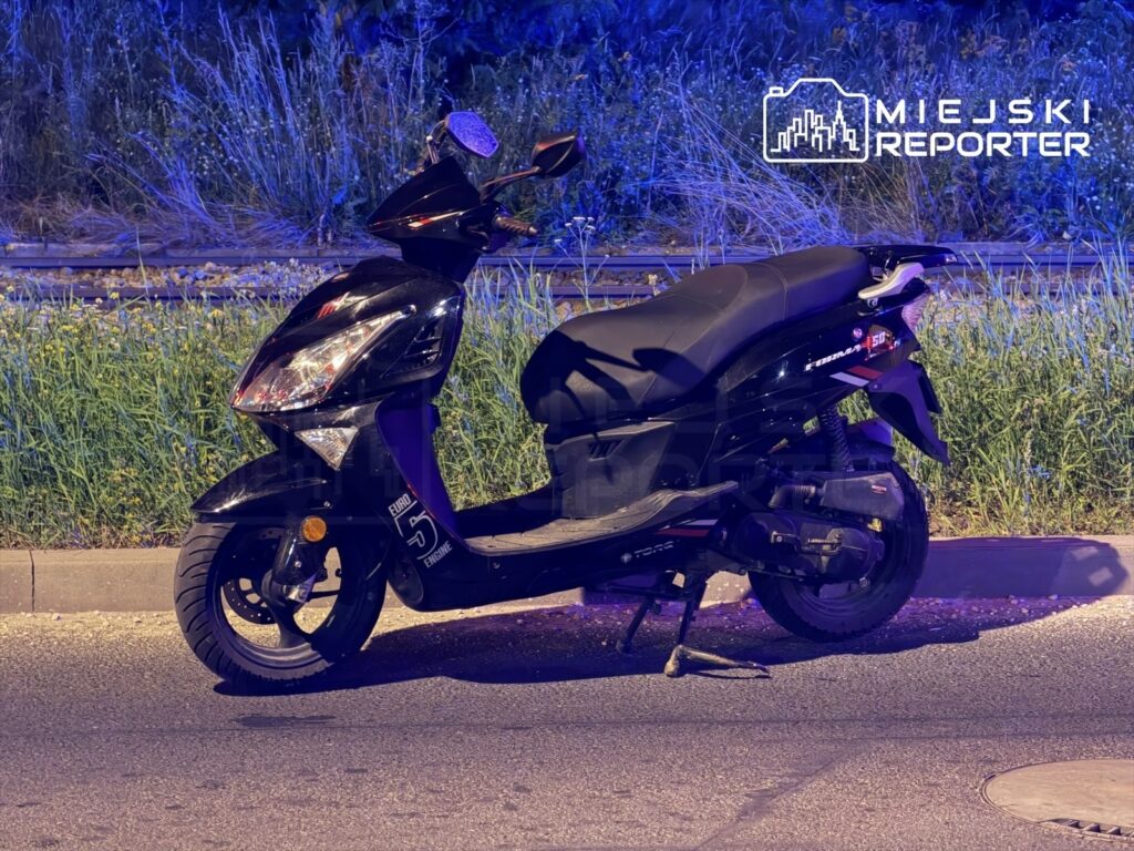 Czarny skuter leży na boku na poboczu drogi, otoczony zielenią i oświetlony niebieskimi światłami.