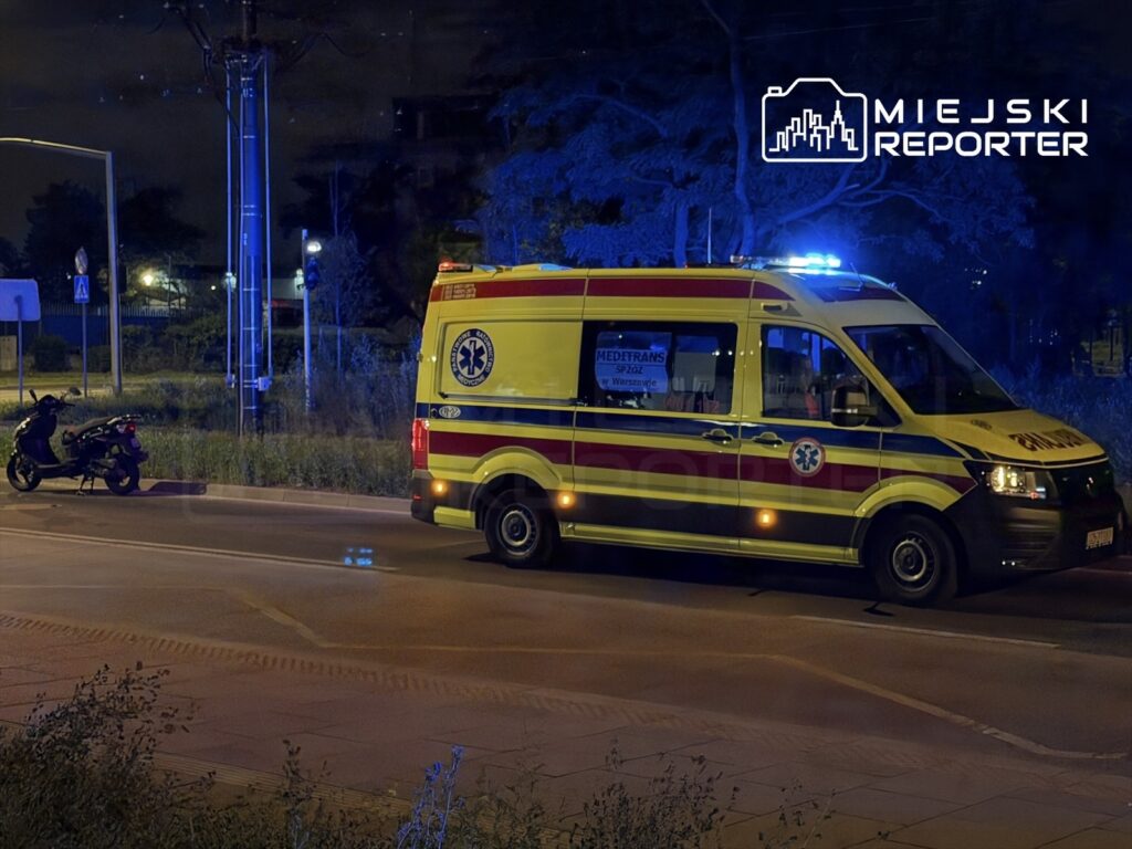 Karetka pogotowia z włączonymi sygnałami świetlnymi zatrzymana obok przewróconego motocykla na oświetlonej ulicy.