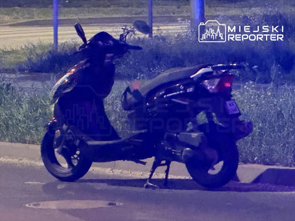 Skuter leży na boku obok krawężnika, w otoczeniu trawy i asfaltowej drogi.