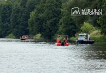 Grupa ratowników na czerwoną łodzią przeszukuje rzekę, obok policyjna motorówka, otoczone zaroślami.