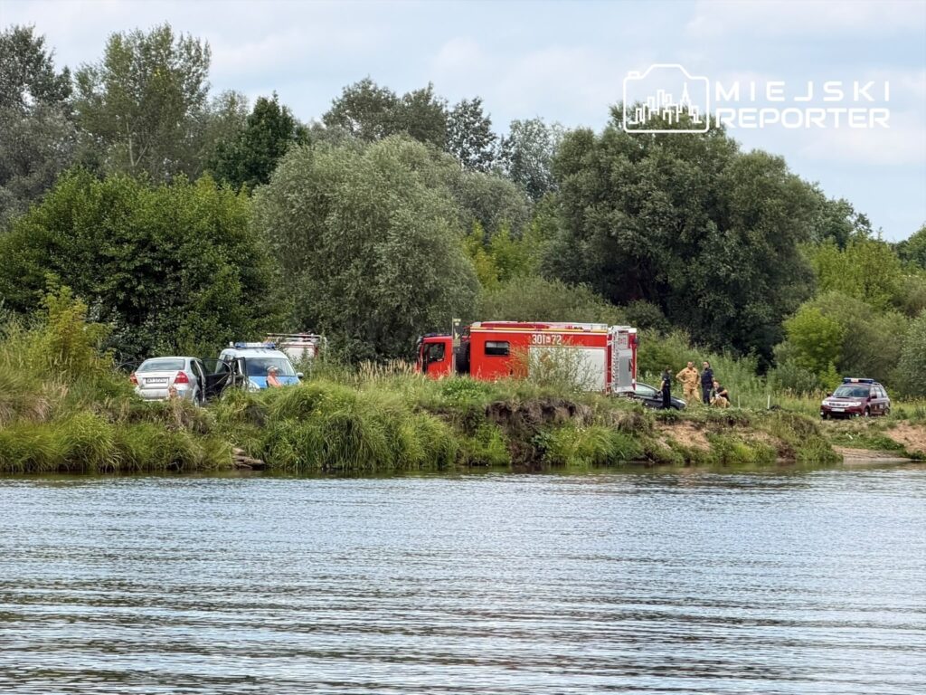 Grupa ratowników pracuje na brzegu rzeki, obok pojazdów służb ratunkowych i osobowych, w otoczeniu zieleni.