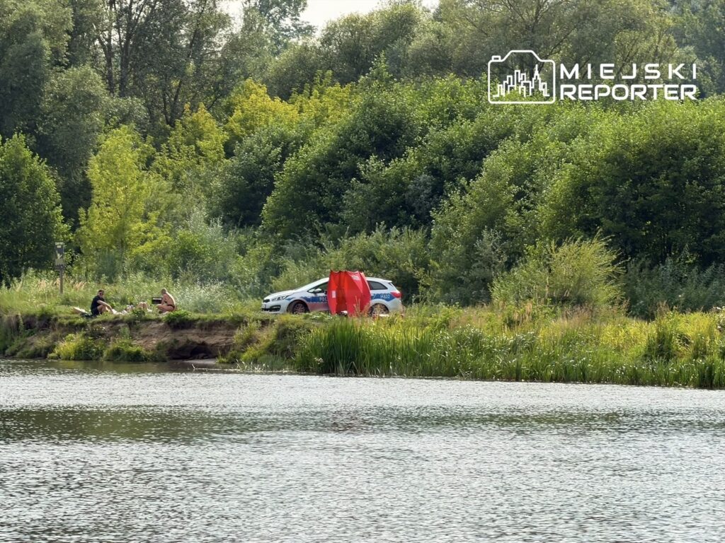 Dwaj mężczyźni siedzą na brzegu rzeki, obok nich leży biały pies, a w tle stoi radiowóz policyjny.