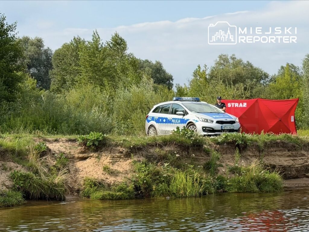 Policjant obok oznakowanego radiowozu stoi na brzegu rzeki, w tle zielona roślinność i czerwony namiot.