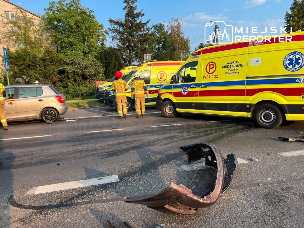 Na ulicy stoją dwa karetki pogotowia, a obok nich pracują strażacy przy uszkodzonym samochodzie. Na jezdni leży zniszczony fragment karoserii.