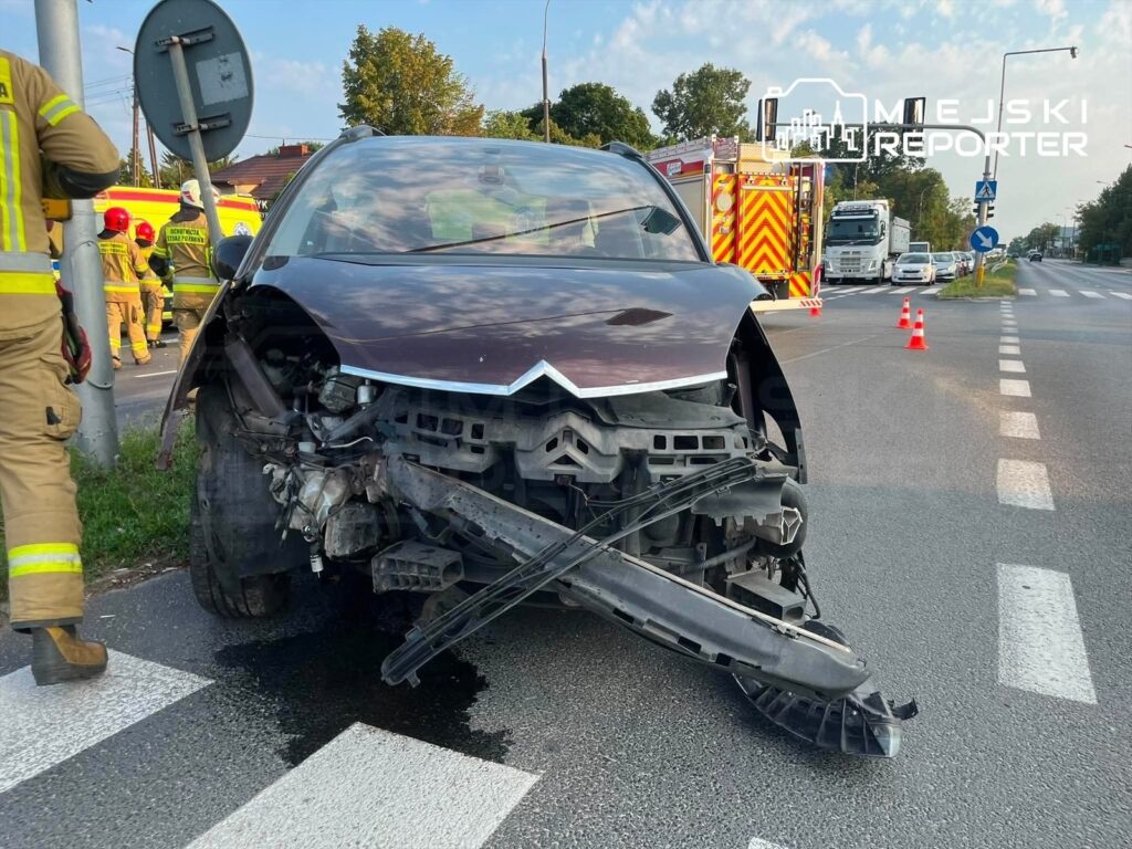 Uszkodzony samochód osobowy z rozbitym przodem stoi na jezdni, obok pracują strażacy oraz pojazdy służb ratunkowych.