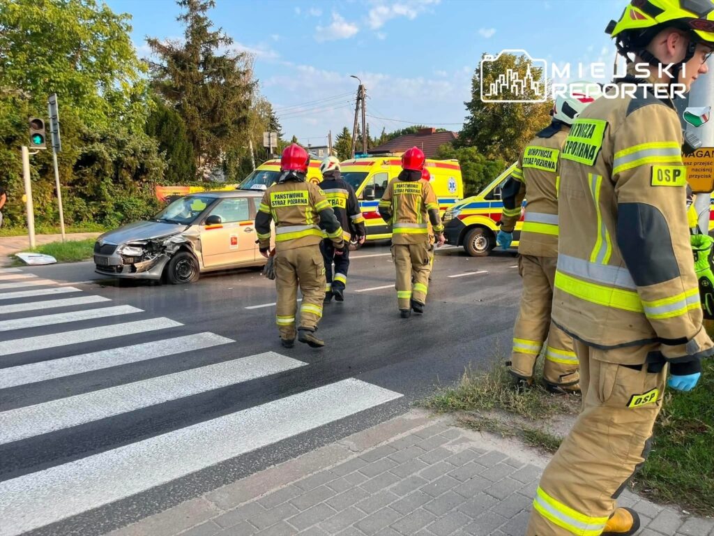 Strażacy w mundurach i kaskach podchodzą do uszkodzonego samochodu na skrzyżowaniu, obok stoją karetki.