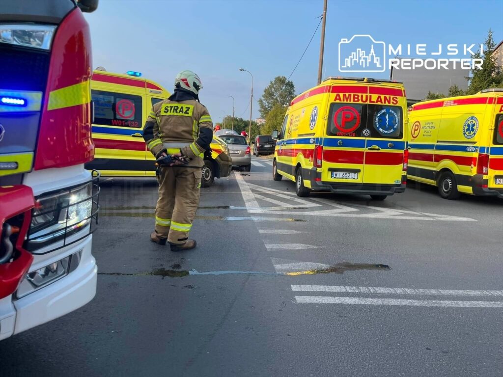 Strażak w mundurze stoi na jezdni obok zablokowanej drogi, a w tle widoczne są karetki pogotowia.