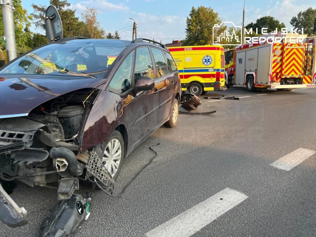 Uszkodzony samochód osobowy z rozbitym przodem stoi na jezdni obok dwóch pojazdów ratunkowych.