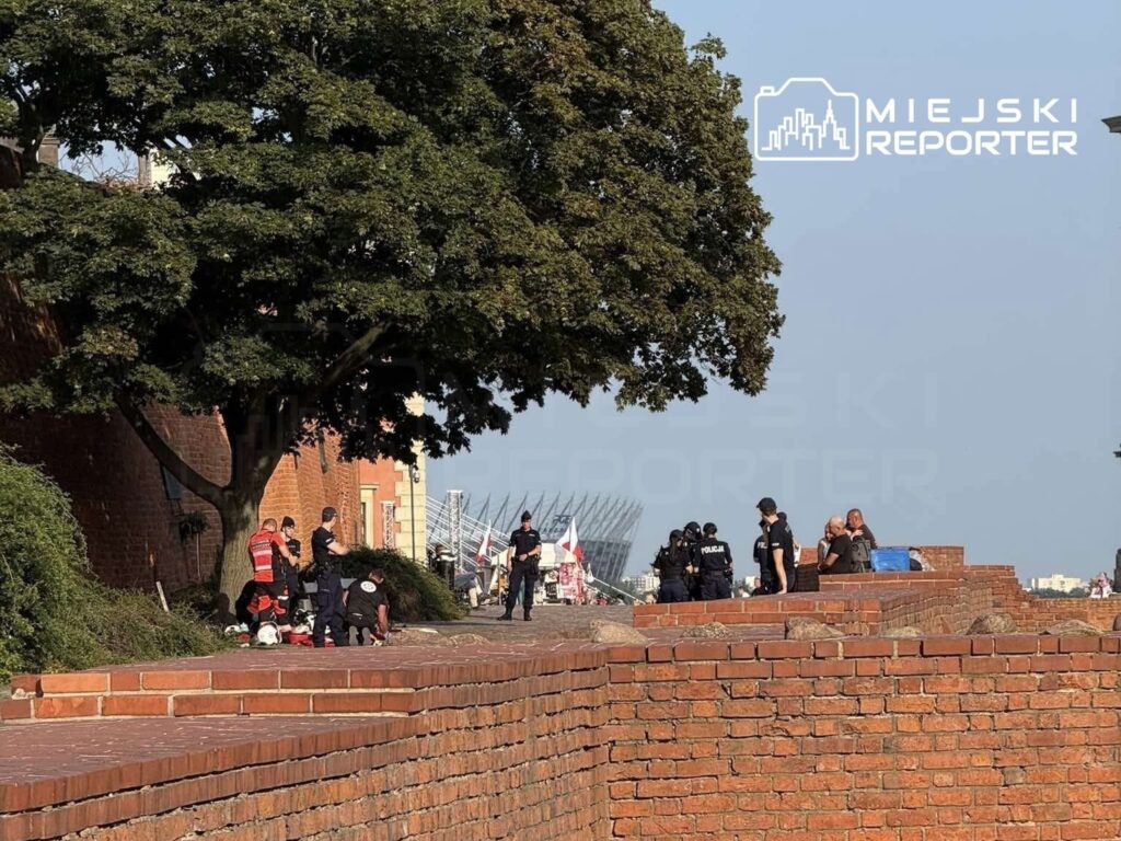 Grupa ratowników i policjantów udziela pomocy na terenie miejskiego murku, w tle widoczny port i flagi.