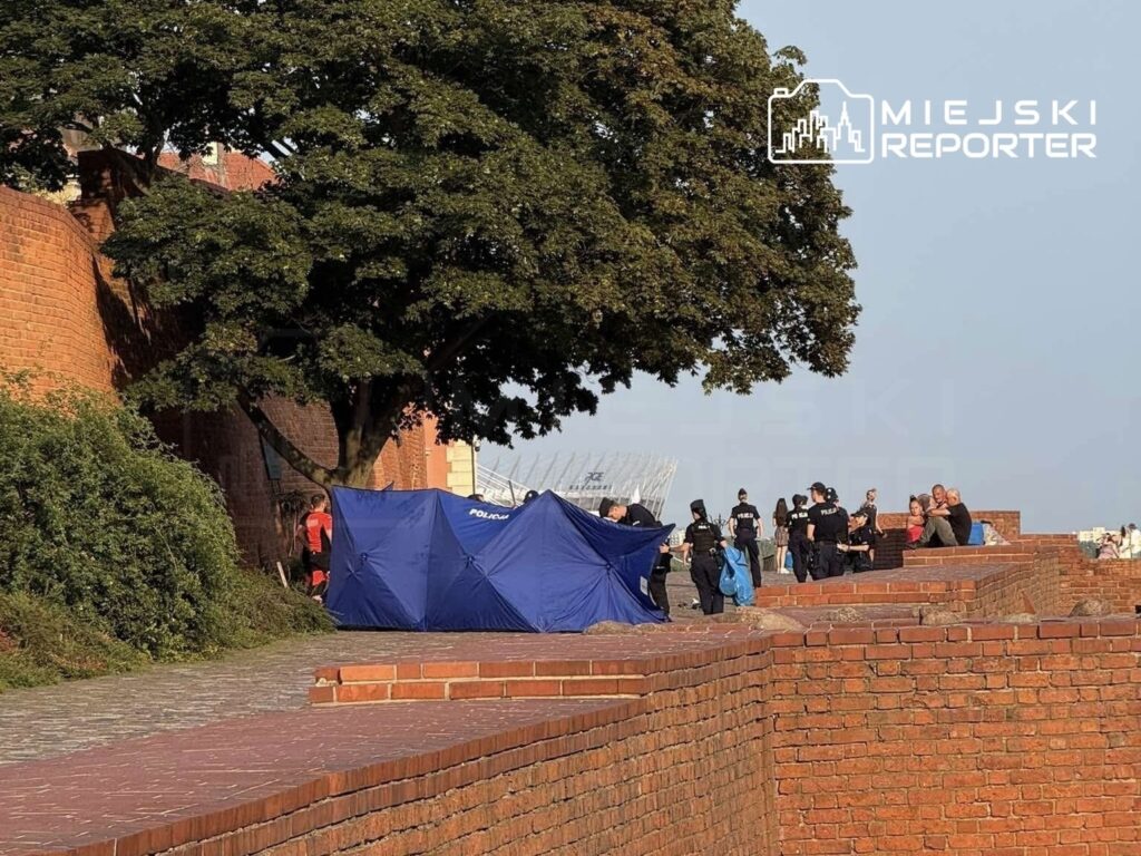 Grupa policjantów w mundurach zbiera się wokół niebieskiego namiotu na tle murów z czerwonej cegły.